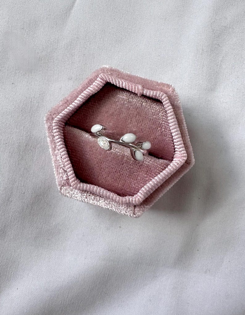 May include: A silver ring with a delicate branch design featuring white enamel leaves and a small, clear gemstone. The ring is displayed inside a pink velvet jewelry box, set against a soft, white background.