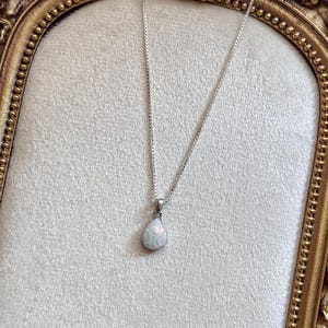 May include: A silver box chain necklace featuring a teardrop-shaped pendant with a milky white opal-like stone that displays iridescent flashes of color. The pendant is securely attached to the delicate metallic chain with a simple silver bail.