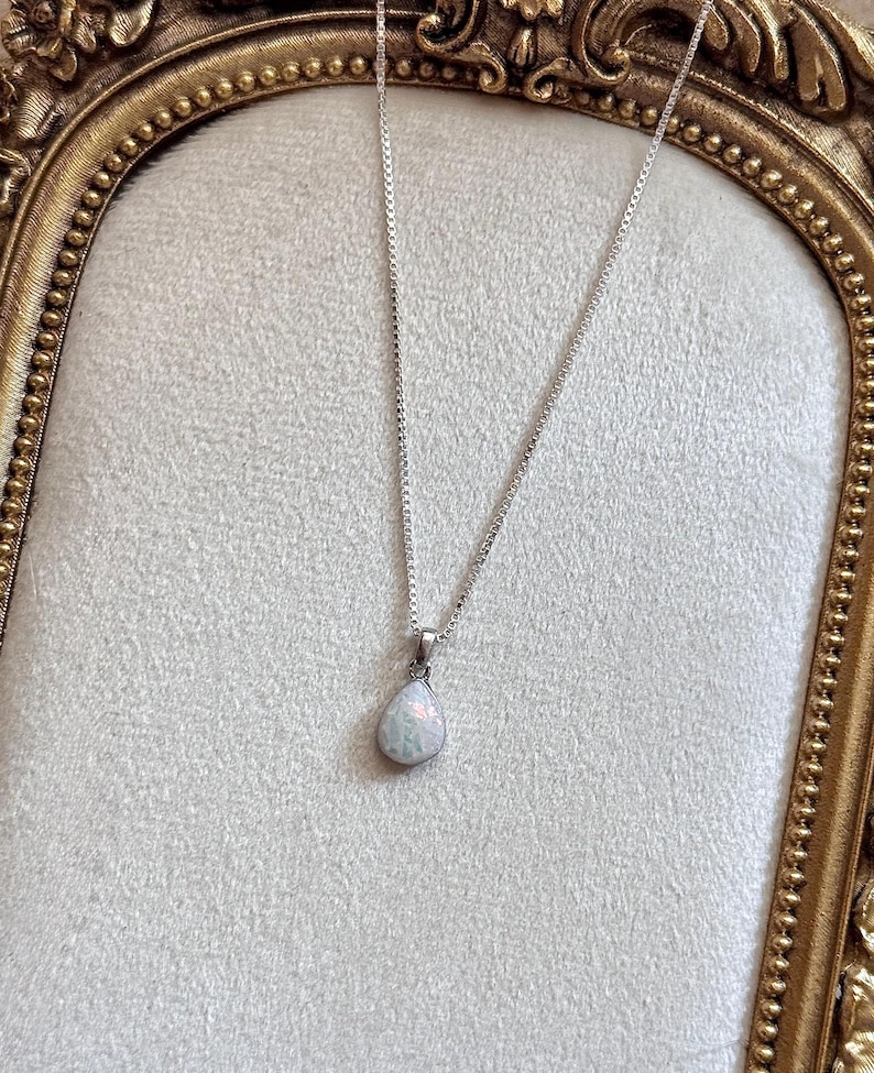 May include: A silver box chain necklace featuring a teardrop-shaped pendant with a milky white opal-like stone that displays iridescent flashes of color. The pendant is securely attached to the delicate metallic chain with a simple silver bail.