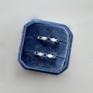 May include: Two silver rings with white and blue gemstones are displayed inside a blue velvet ring box. The rings have a delicate design, with alternating white and blue stones. The ring box is rectangular and has a soft, plush texture.