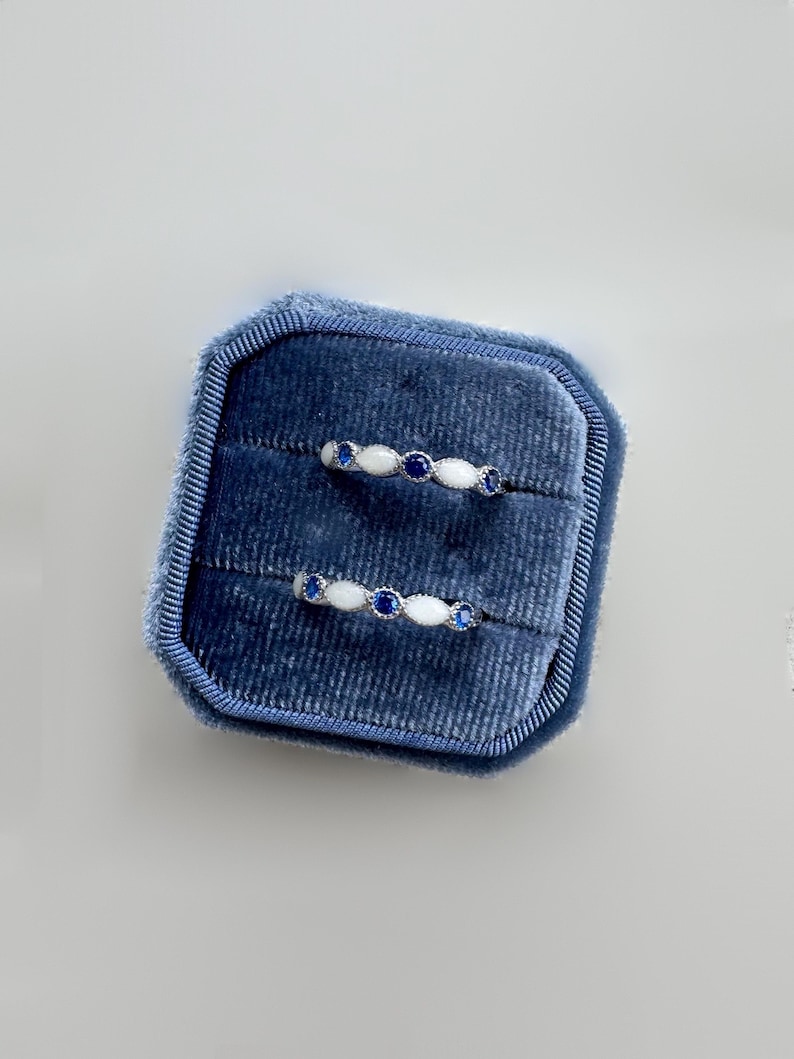 May include: Two silver rings with white and blue gemstones are displayed inside a blue velvet ring box. The rings have a delicate design, with alternating white and blue stones. The ring box is rectangular and has a soft, plush texture.