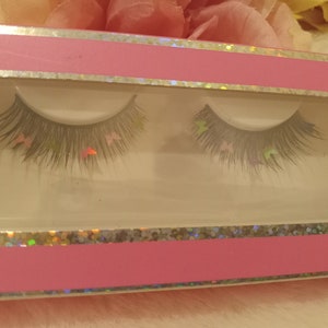 Pode incluir: Um par de cílios postiços com um design de glitter rosa e prata. Os cílios estão em uma caixa de plástico transparente com uma tampa rosa.