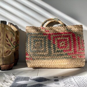 May include: A woven straw tote bag with green and pink geometric patterns. The bag has two handles and is sitting on a white tiled floor.
