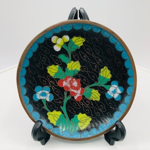 May include: A small, round, black and blue enamel dish with a floral design. The dish has a gold rim and is decorated with a floral pattern in red, yellow, green, and blue.