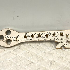 May include: White wooden key-shaped wall decor with gold hooks and handwritten text that reads "Awards Paid at fifty cents / One Hundred Dollars / One Hundred and fifty Dollars / One Hundred Dollars. Be Careful."