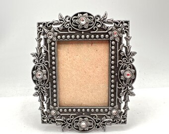 Può includere: Una cornice per foto argentata con un design ornamentale con motivi floreali e strass. La cornice ha un'apertura rettangolare per una foto.