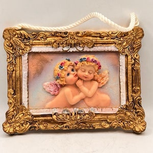 May include: A gold-framed wall decoration featuring two cherubs with floral headbands, one whispering to the other. The cherubs are sculpted in a 3D style and have a soft, pastel colour scheme.