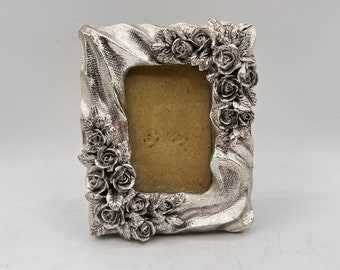 Può includere: Cornice per foto argentata con un design decorativo di rose e foglie. La cornice ha una superficie testurizzata e una forma rettangolare.