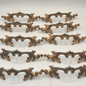 May include: Ten bronze-coloured, ornate drawer pulls arranged in rows. Each pull features a curved design with scrolling leaf and floral details. The hardware is likely for furniture restoration or decorative use.