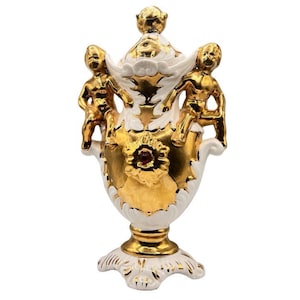 Può includere: Un vaso in ceramica bianca con dettagli dorati e due cherubini sui lati. Il vaso ha un corpo rotondo e una base svasata. I cherubini si aggrappano al vaso e hanno ali dorate. Il vaso ha un gioiello rosso al centro.