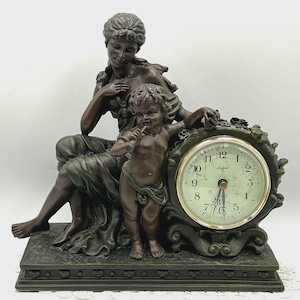Puede incluir: Un reloj de sobremesa color bronce que presenta una figura sentada y un niño de pie tocando la flauta. La esfera es redonda con un borde dorado, números negros y la palabra "Valerie" impresa en el centro. La base es rectangular.