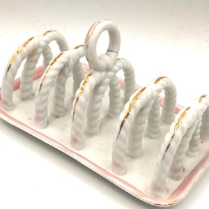 May include: White ceramic toast rack with a pink border and gold accents. The rack features a series of arched supports with a rope-like texture. A handle is at the top. A decorative piece for the dining table.