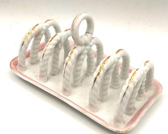 May include: White ceramic toast rack with a pink border and gold accents. The rack features a series of arched supports with a rope-like texture. A handle is at the top. A decorative piece for the dining table.