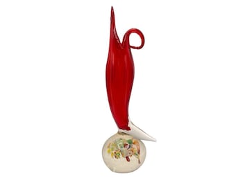 May include: A red glass sculpture with a white base. The sculpture is shaped like a stylized flower with a long, thin stem and a large, round head. The head is decorated with colorful glass pieces.