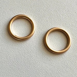 Puede incluir: Dos anillos de metal de color dorado, cada uno con forma circular. Los anillos están colocados planos sobre una superficie blanca texturizada.