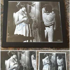 May include: Black and white framed photograph of a vintage movie scene. A woman in a light dress and a man in a button-down shirt are featured. The photograph is signed in blue ink. The frame is black.