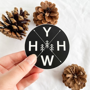 May include: Black and white round sticker with the text "YHHW" and a stylized image of pine trees. The sticker is being held by a hand against a white background.