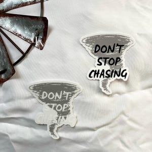 May include: Two stickers with a gray tornado design and the words "DON'T STOP CHASING" in black and white. The stickers are on a white background. A metal windmill is in the upper left corner.