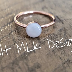 May include: A rose gold ring with a white round gemstone. The ring is on a wooden surface with the text "Spilt MLK Designs" written in white.