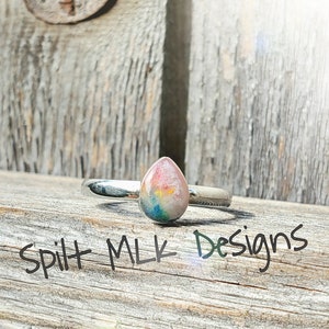 May include: A silver ring with a teardrop-shaped opal gemstone. The opal is a mix of pink, blue, green, and yellow colors. The ring is on a wooden surface with the text "Spilt Milk Designs" written on it.