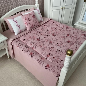 Può includere: Letto bianco in miniatura con copriletto e cuscini a fantasia floreale rosa. La struttura del letto ha una testiera e una pediera bianche con dettagli dorati. Un piccolo comodino bianco con una pianta è accanto al letto.