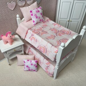 May include: A miniature white bunk bed with pink bedding and pillows. The bedding features a rainbow pattern in shades of pink and orange. Decorative pillows have a white background with pink stars. A small white nightstand holds a pink unicorn.