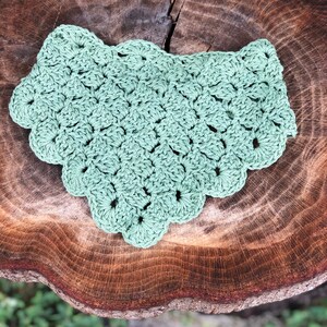 Puede incluir: Un babero de ganchillo verde salvia con un borde festoneado. El babero hecho a mano est&aacute; hecho de un hilo suave y se muestra sobre una superficie de madera r&uacute;stica. La textura y el color del babero sugieren accesorios para beb&eacute;s o artesan&iacute;as.