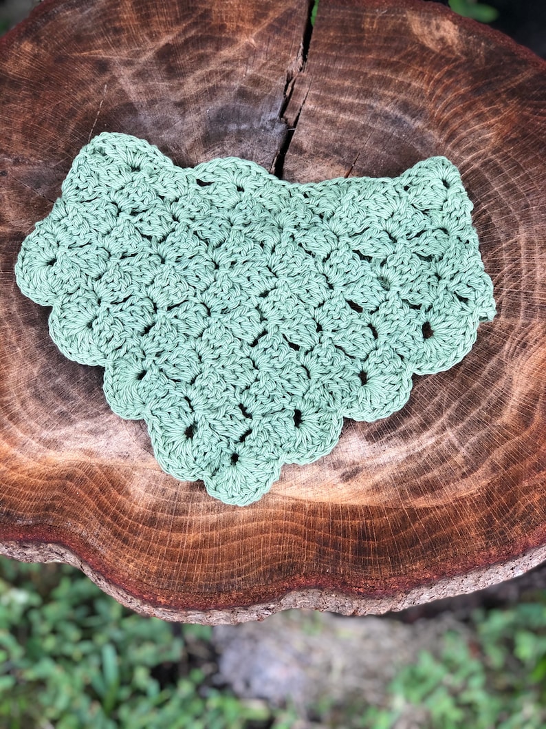 Puede incluir: Un babero de ganchillo verde salvia con un borde festoneado. El babero hecho a mano est&aacute; hecho de un hilo suave y se muestra sobre una superficie de madera r&uacute;stica. La textura y el color del babero sugieren accesorios para beb&eacute;s o artesan&iacute;as.