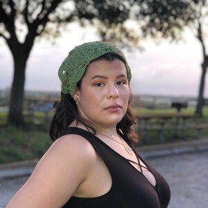 Puede incluir: Se lleva una diadema verde de ganchillo. La diadema est&aacute; hecha de un hilo texturizado y tiene un tejido ligeramente abierto. La persona lleva una camiseta sin mangas negra y un collar. El fondo est&aacute; borroso con &aacute;rboles y un cielo crepuscular.