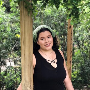 Puede incluir: Una mujer con una diadema verde de ganchillo y un top negro sin mangas. El fondo presenta un exuberante follaje verde y postes de madera, creando un entorno natural al aire libre. La mujer est&aacute; sonriendo.