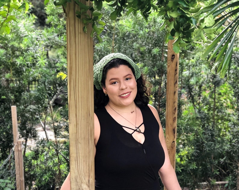 Puede incluir: Una mujer con una diadema verde de ganchillo y un top negro sin mangas. El fondo presenta un exuberante follaje verde y postes de madera, creando un entorno natural al aire libre. La mujer est&aacute; sonriendo.
