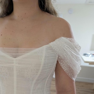 Puede incluir: Cuerpo de encaje blanco con diseño de hombros descubiertos y mangas transparentes adornadas. La parte superior estilo corsé presenta delicados detalles de encaje floral y un ajuste estructurado. Las mangas son de tela transparente con pequeños adornos brillantes.