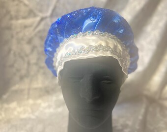 Puede incluir: Un gorro de ducha de satén azul con una banda de satén blanco y ribete plateado. El gorro está en una cabeza de maniquí negra.