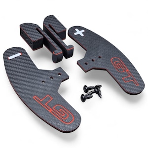 Puede incluir: Un juego de levas de cambio de coche de fibra de carbono negra con detalles en rojo y las letras "GT". Incluye accesorios de montaje. Las levas tienen una superficie texturizada y un símbolo "+" y "-" blanco.