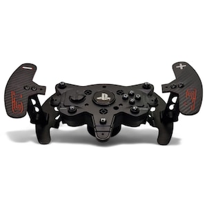 Puede incluir: Volante de carreras negro con levas de cambio de fibra de carbono. El volante presenta un logotipo de PlayStation y varios botones de control. Las levas tienen la marca "GT" roja y símbolos de más/menos. Se ve un botón PS5.