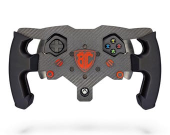 May include: A carbon fiber racing steering wheel controller featuring black ergonomic grips, a central red shield emblem with the letters BC, a directional pad, and four colored action buttons labeled X, Y, A, and B, with an Xbox logo at the base.