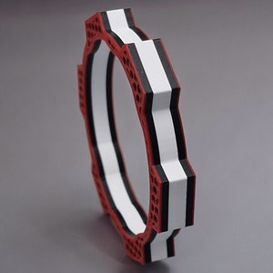 May include: A circular, geometric object with a red, white, and black color scheme. The design features a series of repeating shapes and a honeycomb-like pattern on the red sections. The object is viewed at an angle, highlighting its layered construction.