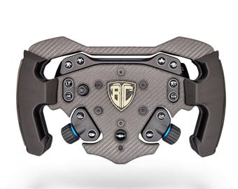May include: A dark gray carbon fiber racing steering wheel controller featuring a central shield emblem with the letters BC. The device includes multiple black buttons labeled with letters and abbreviations, two blue-accented rotary knobs, and ergonomic side grips.