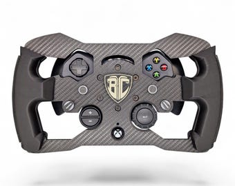 May include: A black carbon fiber racing steering wheel controller featuring a central shield logo with the letters BC. The device includes a directional pad, four colored action buttons, a circular menu dial, a plus-minus button, and an Xbox logo button.