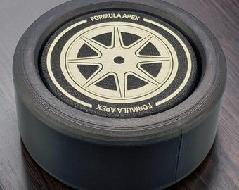 May include: A circular black container with a textured side and a flat top lid. The lid features a beige graphic of an eight-spoke wheel design surrounded by a black border. The text FORMULA APEX is printed in beige on the top and bottom edges.