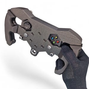 May include: A dark gray carbon fiber textured racing steering wheel controller attachment featuring a directional pad, four colored action buttons labeled A, B, X, and Y, and metallic screw accents. The device has a rigid, angular frame.