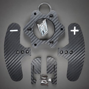 May include: A collection of carbon fiber car parts, including paddle shifters with plus and minus symbols, a central hub with a ring, and mounting hardware. The parts are arranged on a gray surface.