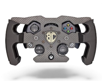 May include: A black carbon fiber racing steering wheel controller featuring a central shield logo with the letters BC, a directional pad, four colored action buttons, a central Xbox button, and two circular control dials on the lower front panel.