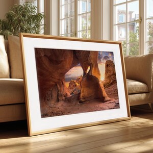 May include: A framed photograph depicting a natural rock archway with a heart-shaped opening, illuminated by the setting sun. The image showcases warm colours and is set in a light wooden frame. The photograph is displayed in a living room, adding a touch of nature to the space.