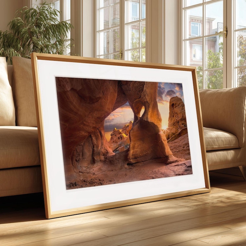 May include: A framed photograph depicting a natural rock archway with a heart-shaped opening, illuminated by the setting sun. The image showcases warm colours and is set in a light wooden frame. The photograph is displayed in a living room, adding a touch of nature to the space.