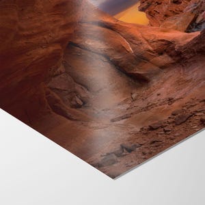 May include: A photograph of a canyon with red rock formations. The image shows a natural archway and a path leading into the distance. The sky is visible in the background with shades of orange and purple.