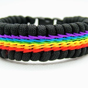 Puede incluir: Una pulsera de paracord negra con un diseño tejido arcoíris. La pulsera tiene un patrón tejido morado, azul, verde, amarillo, naranja y rojo.