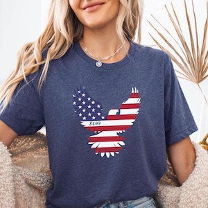 May include: Navy blue t-shirt featuring an eagle design with the American flag incorporated into the wings. The eagle's wings are red and white striped, with a section of blue and white stars. The number "8647" is printed below the eagle.