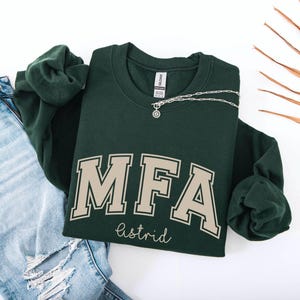 Könnte beinhalten: Ein dunkelgrünes Sweatshirt mit den Buchstaben "MFA" in einer beigefarbenen College-Schriftart, darunter der Name "astrid". Eine silberne Halskette mit Anhänger liegt auf dem Sweatshirt. Das Sweatshirt liegt neben einer hellblauen zerrissenen Jeans.