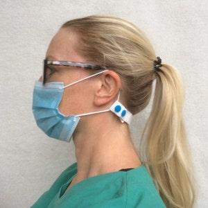 Könnte beinhalten: Eine hellblaue, plissierte Gesichtsmaske mit weißen Ohrschlaufen wird mit einem weißen Riemen und zwei blauen Knöpfen befestigt. Die Maske wird von einer Person mit blonden Haaren und einer Brille getragen.